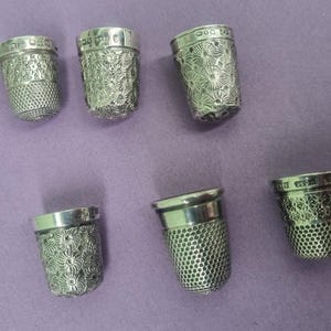 May include: Six silver thimbles with intricate floral designs. The thimbles are all different sizes and shapes, and they are all made of a shiny metal.