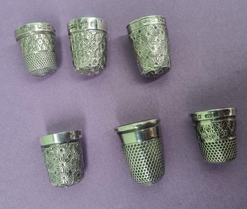May include: Six silver thimbles with intricate floral designs. The thimbles are all different sizes and shapes, and they are all made of a shiny metal.