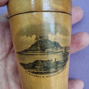 May include: A small wooden box with a hinged lid. The box is decorated with a black and white image of St Michael's Mount, Penzance.