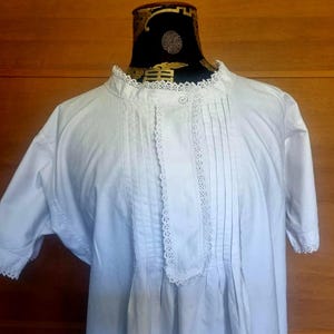 May include: A white vintage nightdress with short sleeves and a high collar. The nightdress features vertical pleats down the front and delicate lace trim along the collar, placket, and sleeve edges. The garment is displayed on a mannequin.