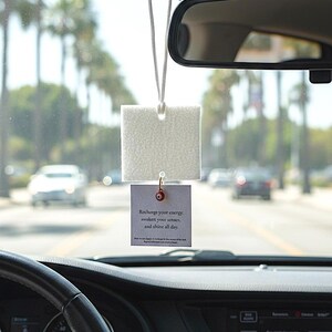 Pode incluir: Um ambientador de carro quadrado branco pendurado por um fio, com um pequeno cartão anexado. O cartão diz: "Recarregue sua energia, desperte seus sentidos e brilhe o dia todo."