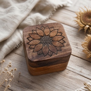 May include: A square wooden box with rounded corners, featuring a dark brown finish and a detailed sunflower design engraved on the lid. The box has a visible wood grain texture and a two-tone appearance with a darker lid and lighter base.