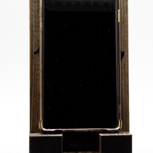 Pode incluir: Um suporte de telefone de madeira com uma mancha marrom escura. Um smartphone preto com uma moldura dourada est&aacute; no suporte.