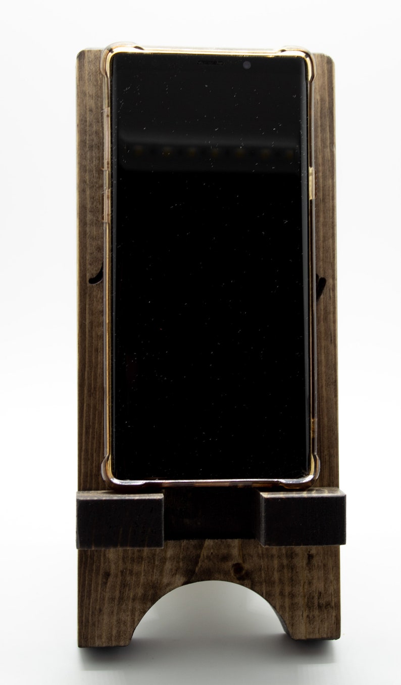 Pode incluir: Um suporte de telefone de madeira com uma mancha marrom escura. Um smartphone preto com uma moldura dourada est&aacute; no suporte.