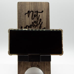 Pode incluir: Um suporte de telefone de madeira com as palavras "Not all who wander are lost" gravadas na frente. Um smartphone preto est&aacute; apoiado no suporte.