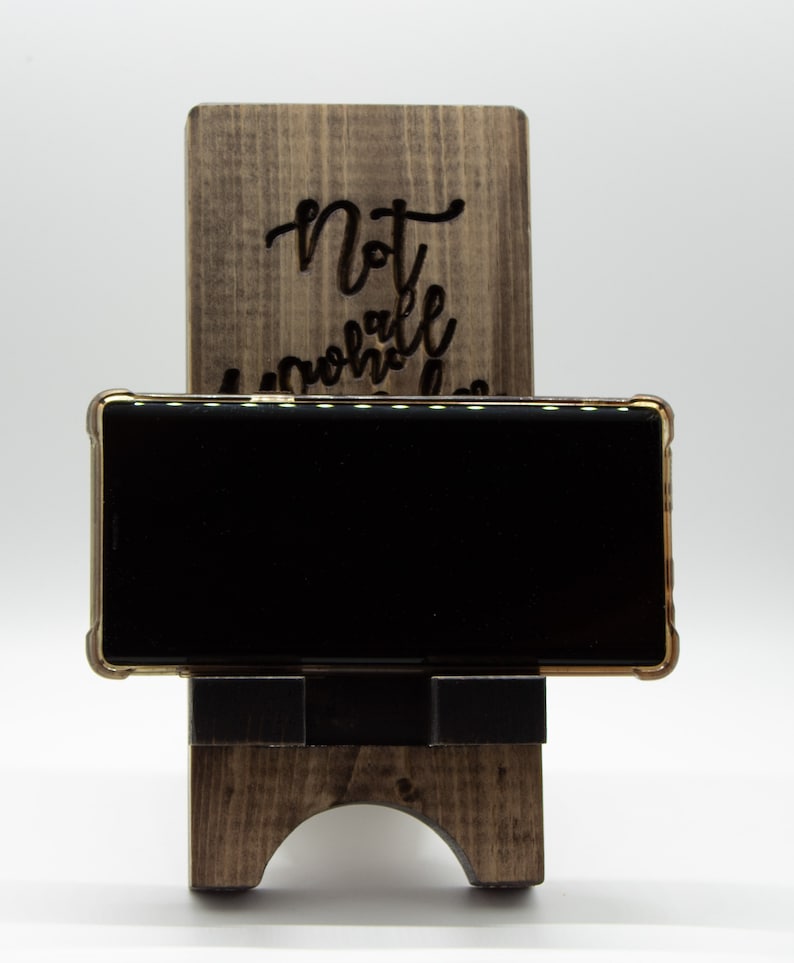 Pode incluir: Um suporte de telefone de madeira com as palavras "Not all who wander are lost" gravadas na frente. Um smartphone preto est&aacute; apoiado no suporte.