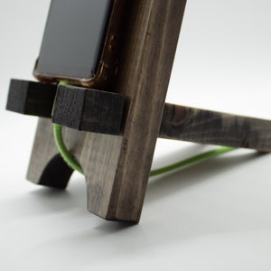 Pode incluir: Um suporte de telefone de madeira marrom escuro com um cord&atilde;o verde passando por ele. O suporte tem dois blocos pequenos nas laterais para segurar o telefone no lugar.