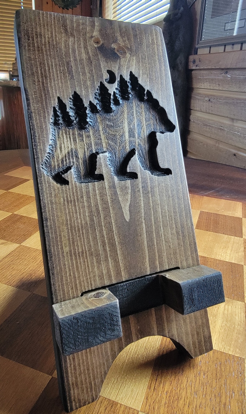 Pode incluir: Um suporte de telefone de madeira com um design entalhado de um urso e &aacute;rvores. O suporte &eacute; marrom escuro com gr&atilde;o de madeira natural. O urso est&aacute; virado para a direita e tem uma lua crescente acima dele.