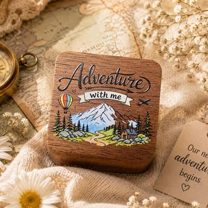 May include: A square wooden box featuring a laser-engraved mountain landscape with a hot air balloon and a small cabin. The text Adventure with me is engraved on the lid. A separate paper tag reads Our next adventure begins with a heart symbol.