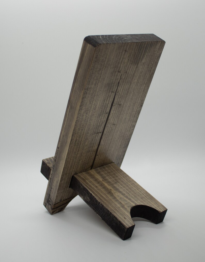 Pode incluir: Um suporte de telefone de madeira com uma mancha marrom escura. O suporte &eacute; feito de duas pe&ccedil;as de madeira, uma para a base e outra para o suporte do telefone. O suporte do telefone &eacute; inclinado para segurar um telefone na vertical.