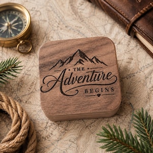 May include: A square wooden box with rounded corners featuring a laser-engraved mountain graphic and the text "THE Adventure BEGINS" with a small heart symbol. The box has a natural brown wood grain finish and a smooth, flat top surface.
