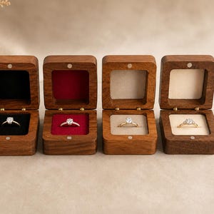 May include: Four square wooden ring boxes with hinged lids, each containing a gold ring with a single clear stone. The interior linings are black, red, beige, and cream, respectively, and each box features a small magnetic closure.