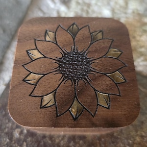 May include: A wooden box with a sunflower design carved into the top. The sunflower is outlined in black and has gold accents.