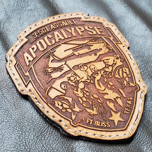 May include: A brown leather patch with the words "APOCALYPSE" and "3-501 ASSAULT" in a shield shape. The patch features an engraved design of figures and a star, with "FT BLISS TEXAS" at the bottom. White stitching outlines the patch.