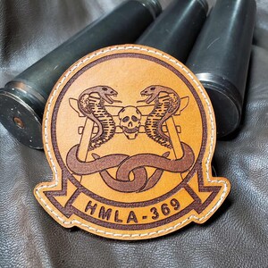 May include: A tan leather patch with a detailed design featuring two cobras, a skull, and the text "HMLA-369". The patch is stitched with white thread and set against a dark background with three dark cylinders.