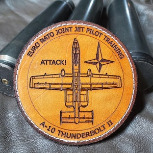 May include: A round, tan leather patch with the text "EURO NATO JOINT JET PILOT TRAINING" and "ATTACK!" and an illustration of an A-10 Thunderbolt II aircraft. The patch is stitched with white thread. Three dark gray cylinders are in the background.
