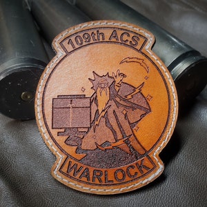 May include: A brown leather patch with the words "109th ACS" and "WARLOCK" in a circular design. The patch features an illustration of a wizard with lightning bolts. The patch is stitched with white thread.