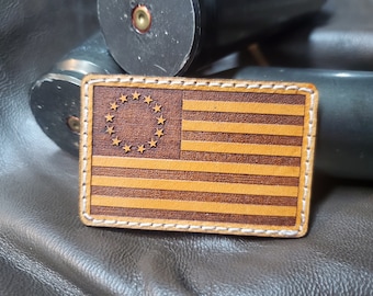 Könnte beinhalten: Ein rechteckiger Lederaufnäher mit dem Design der Betsy-Ross-Flagge. Die Flagge hat 13 Sterne in einem Kreis und 13 Streifen. Der Aufnäher ist braun und hellbraun mit weißen Nähten an den Rändern.