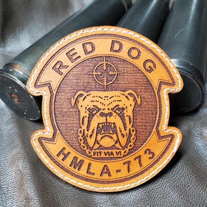May include: A circular leather patch with the words "RED DOG" and "HMLA-773" in a banner design. The patch features a bulldog graphic and a crosshair symbol. The patch is tan with white stitching and is set against a dark background.