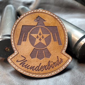 May include: A brown leather patch with the text "Thunderbirds" in cursive script. The patch features a stylized eagle emblem with a star and three small aircraft. The patch is stitched with white thread.