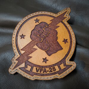 Pode incluir: Um patch de couro bronzeado com um punho marrom segurando um raio, cercado por um círculo e estrelas. O texto "VFA-25" está na parte inferior. O patch tem uma borda costurada.