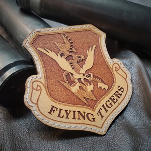 May include: A leather patch with the words "FLYING TIGERS" and a detailed emblem featuring a stylized tiger and lightning bolts. The patch is tan with a brown design and white stitching around the edges.