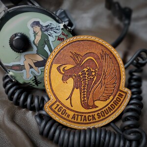 May include: A leather patch with a cobra design and the text "160th Attack Squadron" is displayed. A vintage headset with a pin-up girl sticker is in the background. The patch is brown and yellow with white stitching.