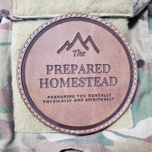 May include: A round, brown leather patch with the text "The Prepared Homestead" and a mountain graphic. The patch is stitched with white thread and is set against a camouflage background. The text below reads "Preparing you mentally, physically, and spiritually."