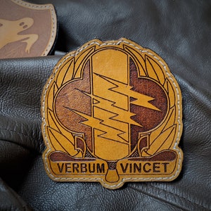 May include: A tan leather patch with a central design of three lightning bolts and the words "VERBUM VINCET". The patch is stitched with white thread and has a decorative border. Another patch with a ghost design is visible in the background.
