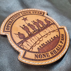 May include: A brown leather patch with white stitching, featuring the text "OPERATION LONE STAR" and "NONE SHALL PASS". The patch depicts silhouettes of soldiers, a star, and barbed wire.