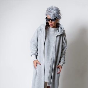 Könnte beinhalten: Ein langer, grauer Kapuzenpullover mit Reißverschluss. Der Pullover hat eine lockere Passform und einen leicht abgenutzten Look. Das Model trägt den Pullover mit einem Paar roten Sneakers.