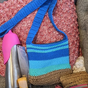 May include: A blue crocheted bag with a brown bottom and a striped design in shades of blue and green. The bag has a long blue strap and is sitting on a couch with a pink water bottle and a roll of yellow tape.