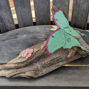 May include: A crocheted green and pink luna moth with a pink flower on a piece of driftwood.