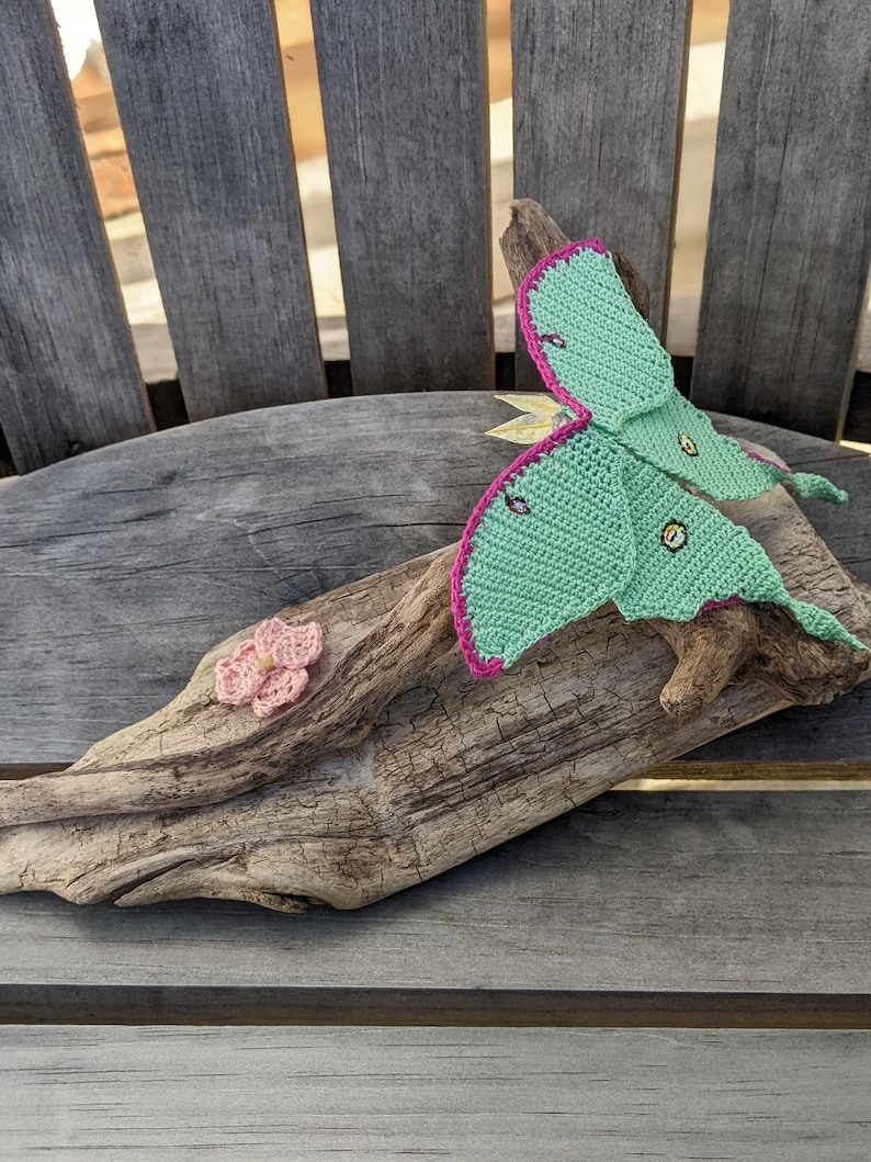 May include: A crocheted green and pink luna moth with a pink flower on a piece of driftwood.