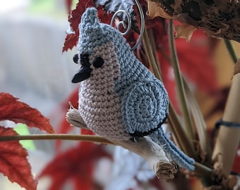 Op de afbeelding: Een gehaakte blauwe vogel ornament met een roze borst en zwarte ogen. De vogel zit op een tak met een draadlus om op te hangen.