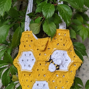 May include: A crocheted tote bag shaped like a honeycomb with a bee applique. The bag is yellow and white with a white strap.