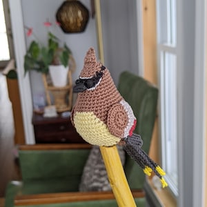 Op de afbeelding: Een gehaakte vogel met bruine, gele en rode accenten, die op een houten stok zit. De vogel heeft zwarte ogen en een zwarte snavel.