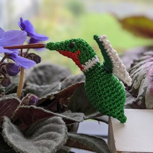 Op de afbeelding: Een gehaakte kolibrie met groene en witte veren, een rode keel en zwarte ogen, die op een wit oppervlak zit, met een paarse bloem op de achtergrond.