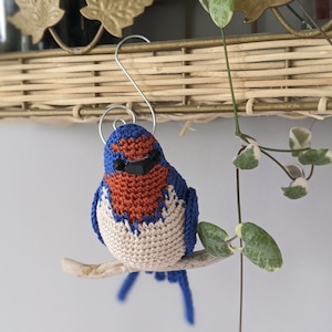 May include: A crocheted bluebird ornament with orange and white accents, perched on a small branch. The bird has black eyes and a small, hooked beak.