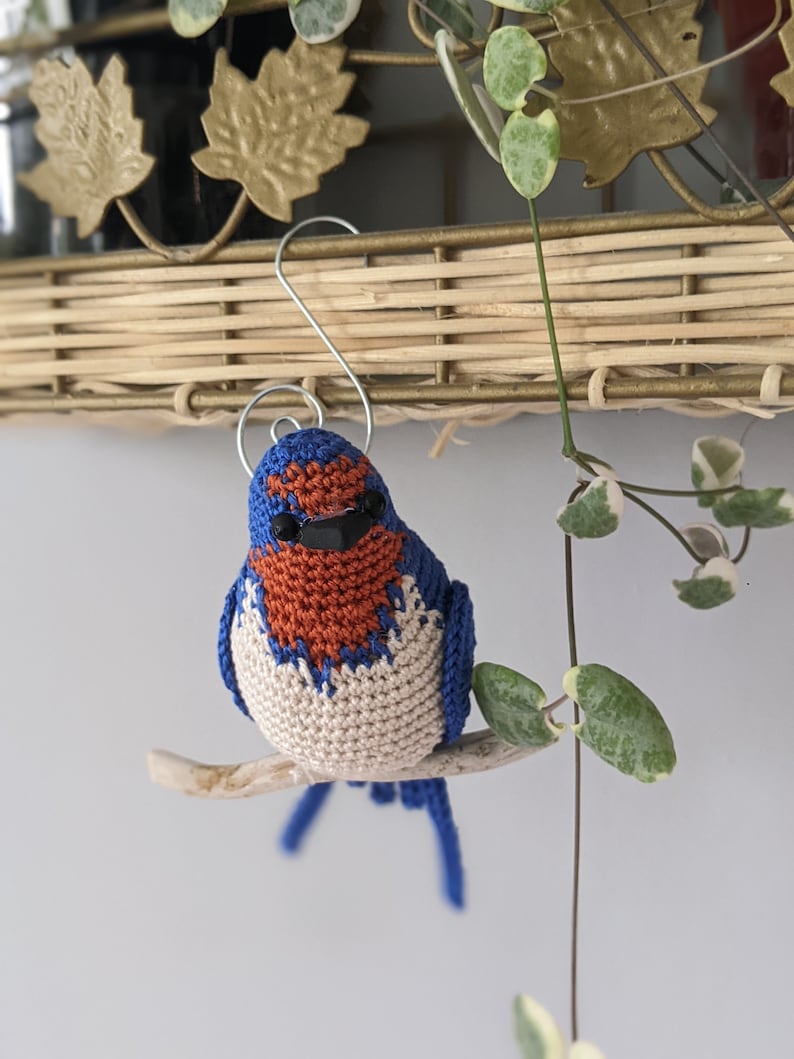 May include: A crocheted bluebird ornament with orange and white accents, perched on a small branch. The bird has black eyes and a small, hooked beak.