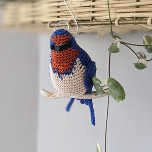 Op de afbeelding: Een gehaakte blauwe vogelversiering met een bruine borst en een zwarte zonnebril. De vogel zit op een tak en hangt aan een haak.
