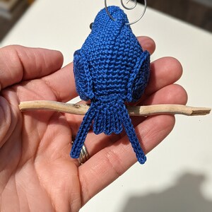 May include: A blue crocheted bird ornament with a wire loop for hanging. The bird is perched on a small branch.
