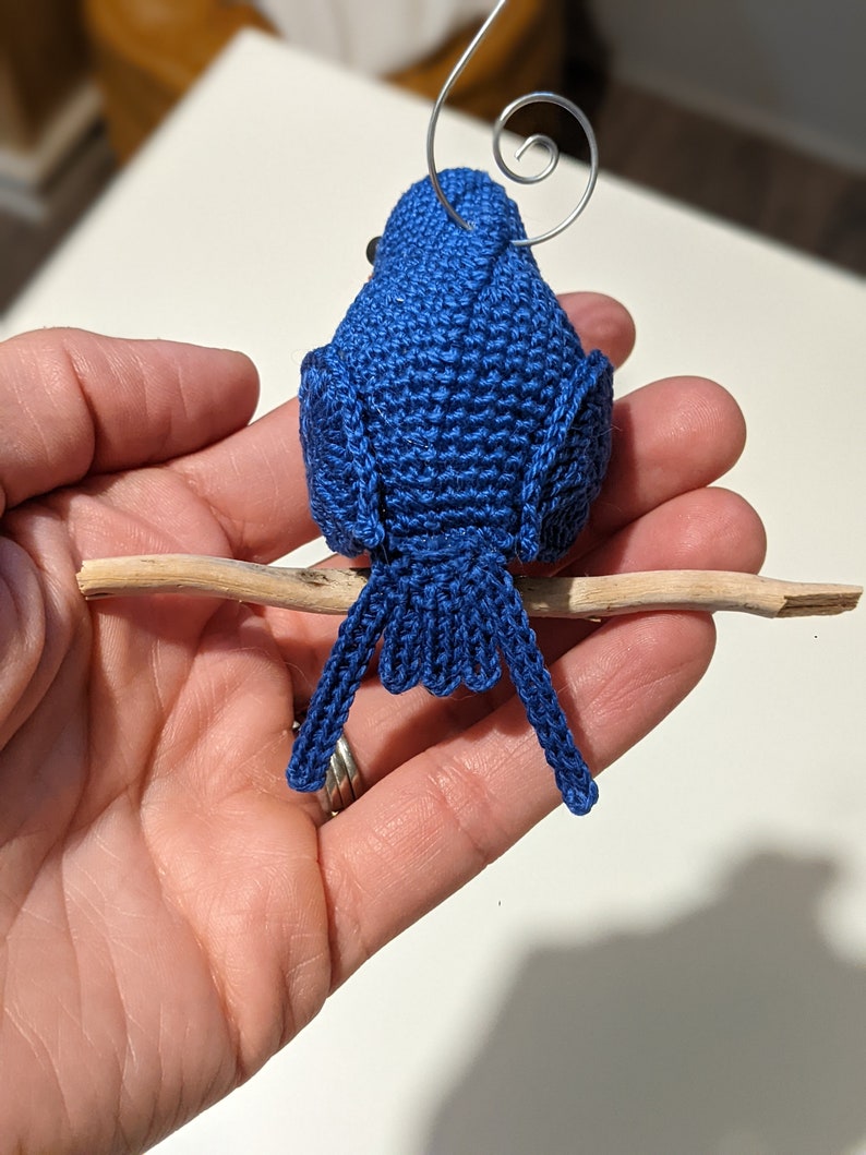 May include: A blue crocheted bird ornament with a wire loop for hanging. The bird is perched on a small branch.