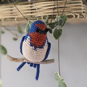 May include: A crocheted bluebird with a brown and white chest, perched on a branch. The bird has black eyes and a small beak.