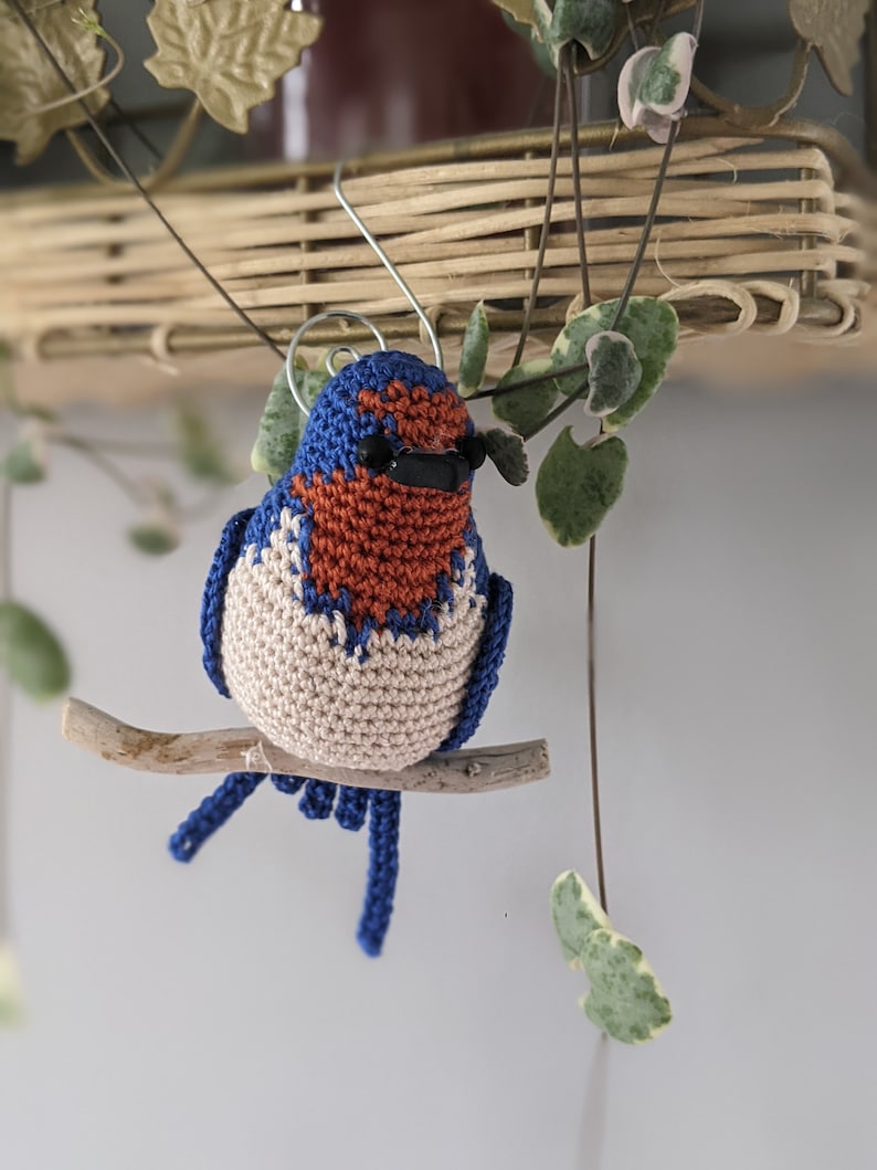 May include: A crocheted bluebird with a brown and white chest, perched on a branch. The bird has black eyes and a small beak.