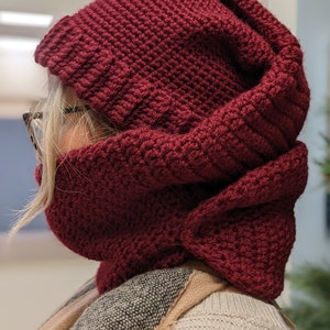 May include: A burgundy crocheted hooded scarf with a thick, textured design. The scarf is folded down to show the hood.