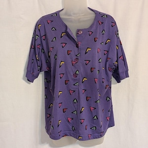May include: A short-sleeved purple shirt featuring a repeating pattern of small, multi-colored triangles in yellow, pink, and green. The shirt has a round neckline with a partial button-down placket containing three matching purple buttons.