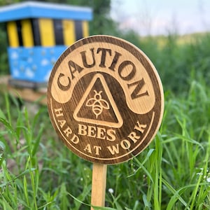Può includere: Un cartello rotondo in legno con la scritta incisa "CAUTION HARD BEES AT WORK" e un simbolo di ape. Il cartello è piantato nell'erba, con un'arnia blu e gialla sullo sfondo.