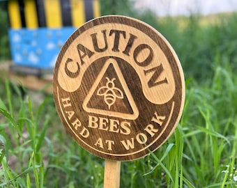 Puede incluir: Un letrero redondo de madera con la inscripción grabada "CAUTION HARD BEES AT WORK" y un símbolo de abeja. El letrero está clavado en la hierba, con una colmena azul y amarilla al fondo.