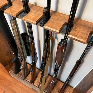 May include: A wooden gun rack displaying several rifles and shotguns. The rack is constructed from stained wood and features slots designed to securely hold the firearms. Various rifles and shotguns are visible, showcasing diverse designs and finishes. The rack is mounted on a wall.